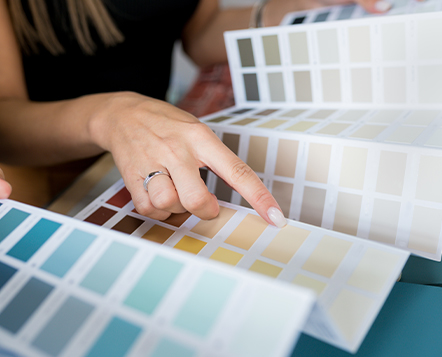 A person with a silver ring on their finger points at a color swatch card, surrounded by various palettes featuring shades of blue, beige, and neutral colors.