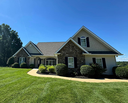 A single-story house with stone and siding exterior, black shutters, and multiple gables sits on a well-manicured lawn under a clear blue sky. Shrubs are neatly arranged along the front walkway.