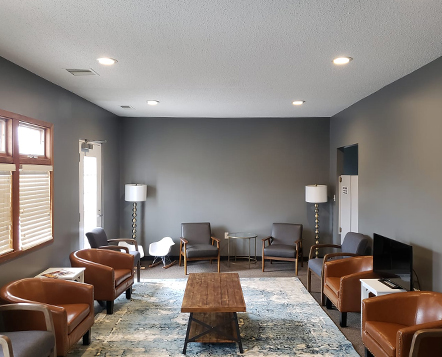 A modern waiting room with brown leather chairs, gray armchairs, a wooden coffee table, two floor lamps, a small TV, and a blue patterned rug, with gray walls and large windows letting in natural light.